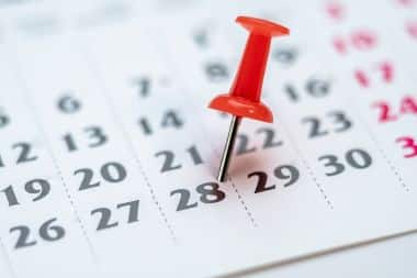 A red push pin is stuck into a paper calendar on the 28th of the month.